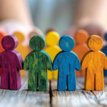 Bonecos coloridos representando diversidade e inclusão, alinhados sobre mesa de madeira enquanto mãos humanas os organizam ao fundo.